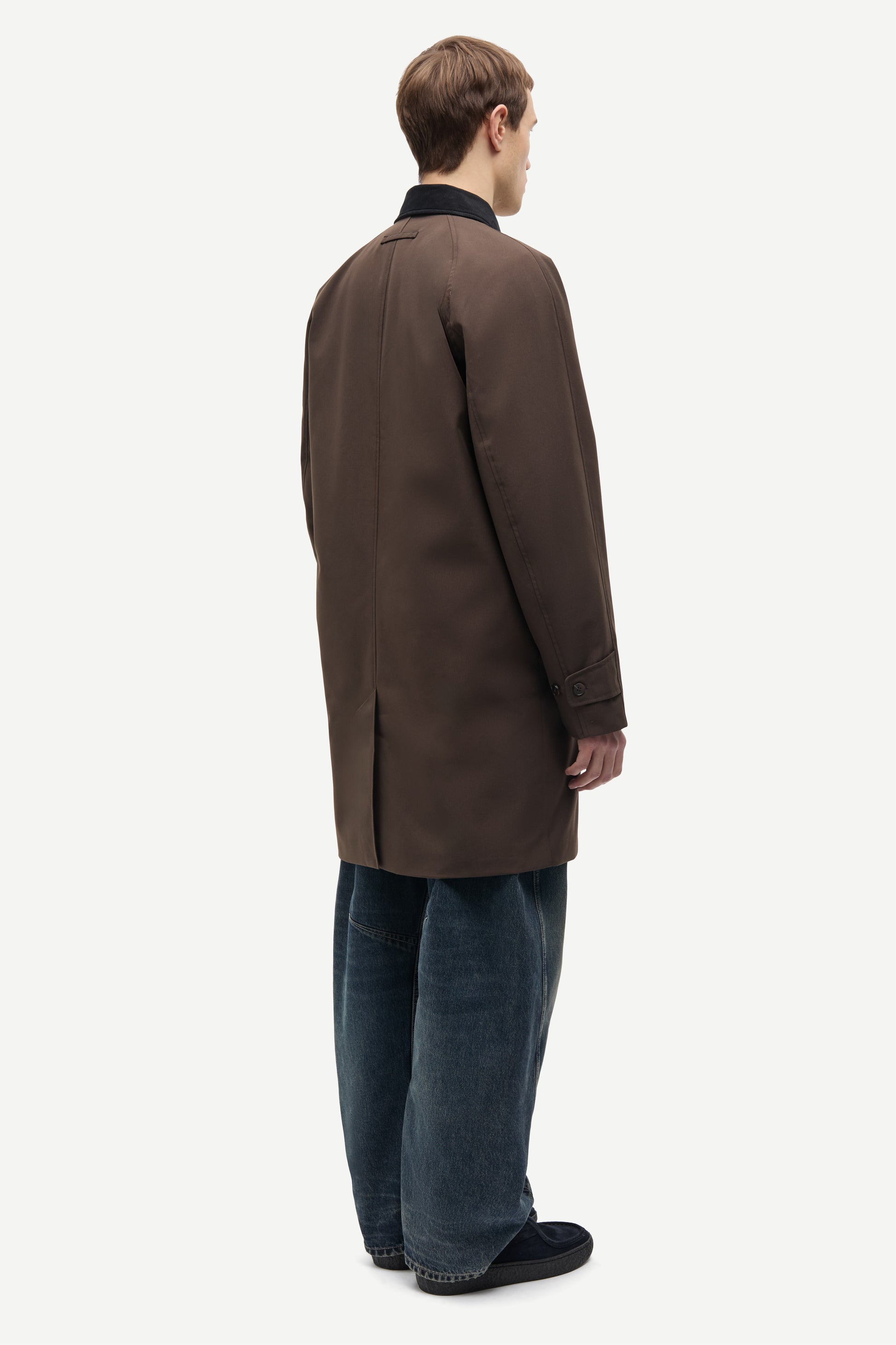 A man shown from the back wearing a dark brown mid-length coat over a black collared shirt, blue jeans, and black slip-on shoes, photographed against a plain light gray studio background.
