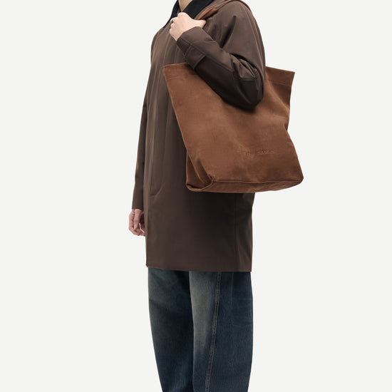 A man in a brown coat and blue jeans stands in profile holding a large brown canvas tote bag over his shoulder, photographed against a plain light gray studio background.