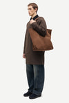 A man in a brown coat and blue jeans stands in profile holding a large brown canvas tote bag over his shoulder, photographed against a plain light gray studio background.