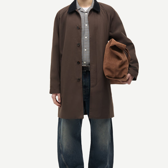 A male model stands against a light gray studio background wearing a dark brown mid-length coat over a checkered shirt and dark blue jeans, while holding a brown suede-like pouch or bag.