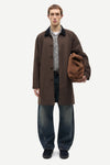 A male model stands against a light gray studio background wearing a dark brown mid-length coat over a checkered shirt and dark blue jeans, while holding a brown suede-like pouch or bag.