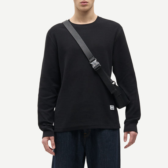 A man wearing a black long-sleeve crewneck sweatshirt and dark blue jeans, carrying a black crossbody bag with a gray strap across his chest, against a light gray studio background.