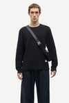 A man wearing a black long-sleeve crewneck sweatshirt and dark blue jeans, carrying a black crossbody bag with a gray strap across his chest, against a light gray studio background.