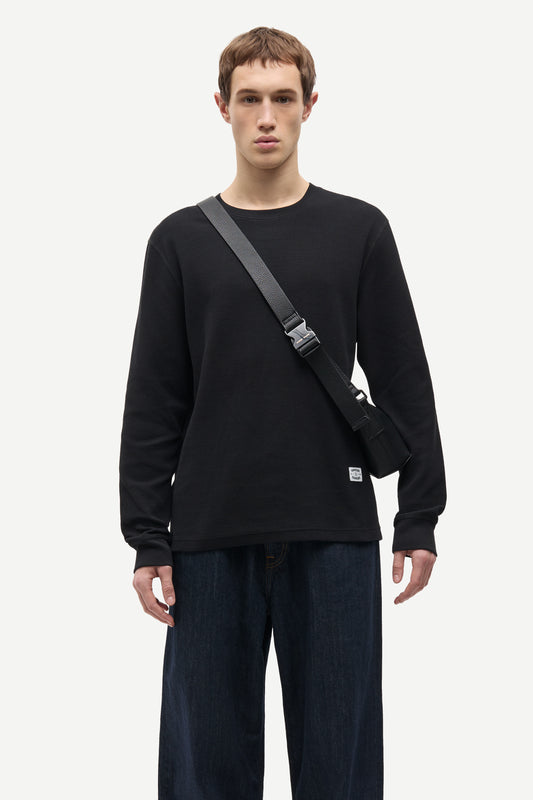 A man wearing a black long-sleeve crewneck sweatshirt and dark blue jeans, carrying a black crossbody bag with a gray strap across his chest, against a light gray studio background.
