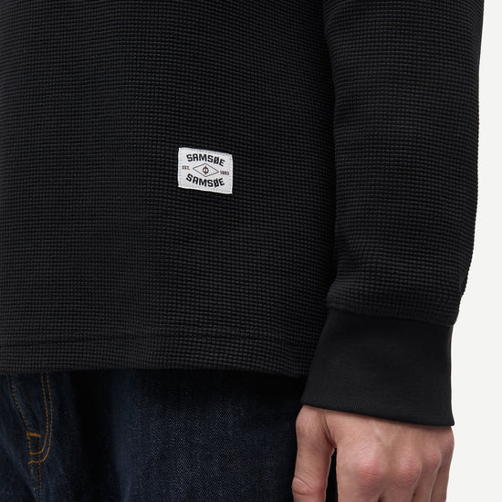 Close-up of a person wearing a black waffle-knit long-sleeve shirt with a white rectangular 'Sakramento' brand patch on the lower left chest, paired with dark blue jeans against a plain light background.