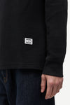 Close-up of a person wearing a black waffle-knit long-sleeve shirt with a white rectangular 'Sakramento' brand patch on the lower left chest, paired with dark blue jeans against a plain light background.