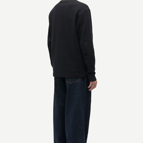 A man shown from the back wearing a black long-sleeve crewneck sweatshirt, dark blue jeans with cuffed hems, and dark green slippers, against a plain light gray background.