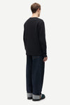 A man shown from the back wearing a black long-sleeve crewneck sweatshirt, dark blue jeans with cuffed hems, and dark green slippers, against a plain light gray background.
