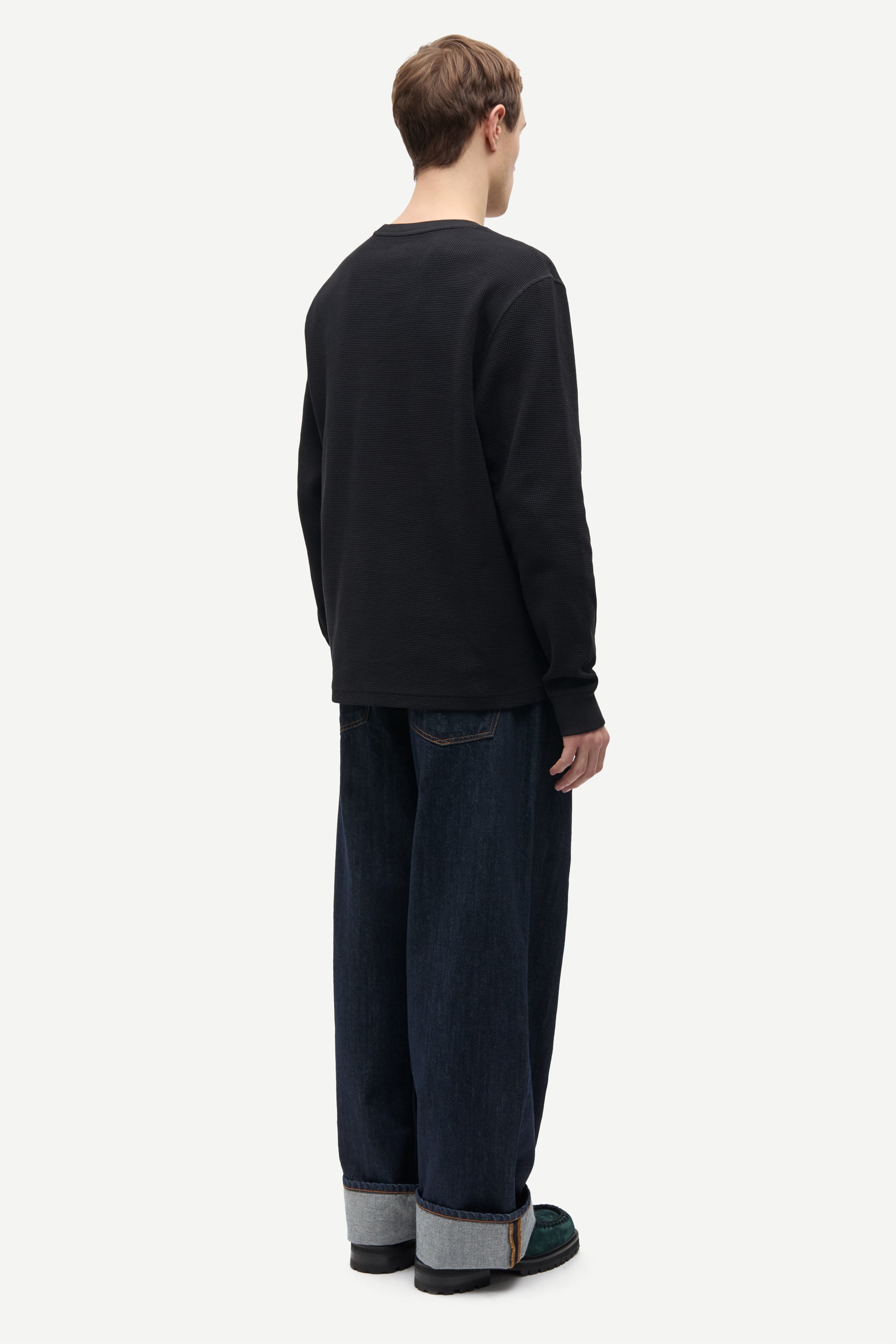A man shown from the back wearing a black long-sleeve crewneck sweatshirt, dark blue jeans with cuffed hems, and dark green slippers, against a plain light gray background.