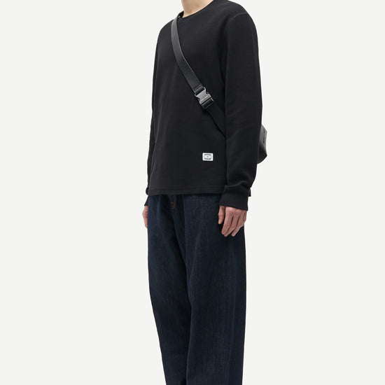 A man stands in profile wearing a black long-sleeve crewneck sweatshirt, dark blue wide-leg jeans with rolled cuffs, dark green slippers, and a black shoulder bag against a plain light gray background.