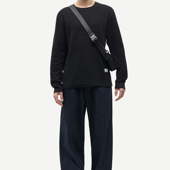 A male model stands against a light gray studio background wearing a black long-sleeve crewneck shirt, dark wide-leg jeans with cuffed hems, dark green shoes, and a black crossbody bag.