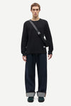A male model stands against a light gray studio background wearing a black long-sleeve crewneck shirt, dark wide-leg jeans with cuffed hems, dark green shoes, and a black crossbody bag.