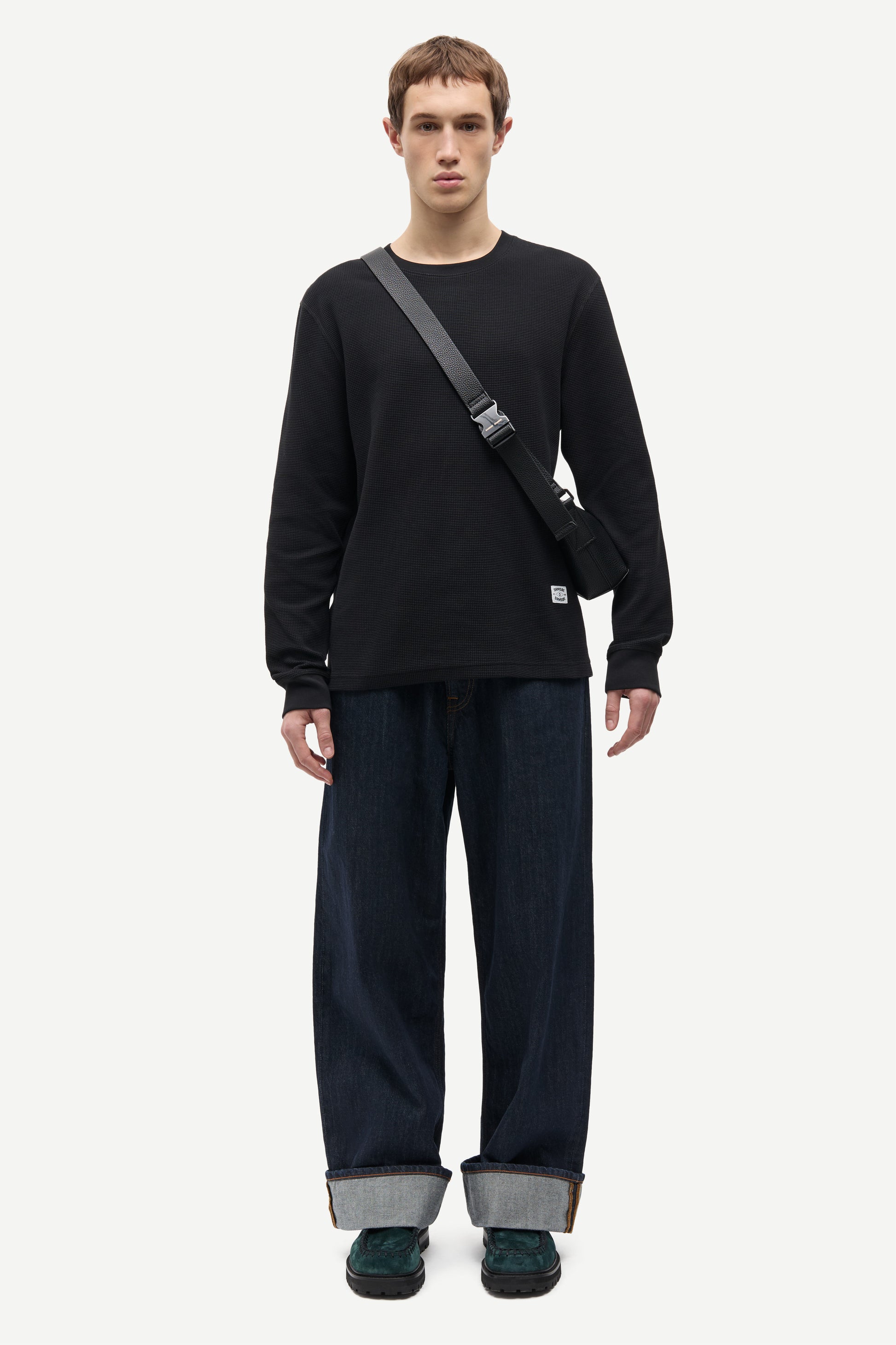 A male model stands against a light gray studio background wearing a black long-sleeve crewneck shirt, dark wide-leg jeans with cuffed hems, dark green shoes, and a black crossbody bag.