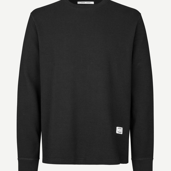 A black long-sleeve crewneck sweatshirt with a small white square logo patch on the lower left hem, displayed on a clean white background.