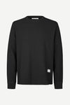 A black long-sleeve crewneck sweatshirt with a small white square logo patch on the lower left hem, displayed on a clean white background.