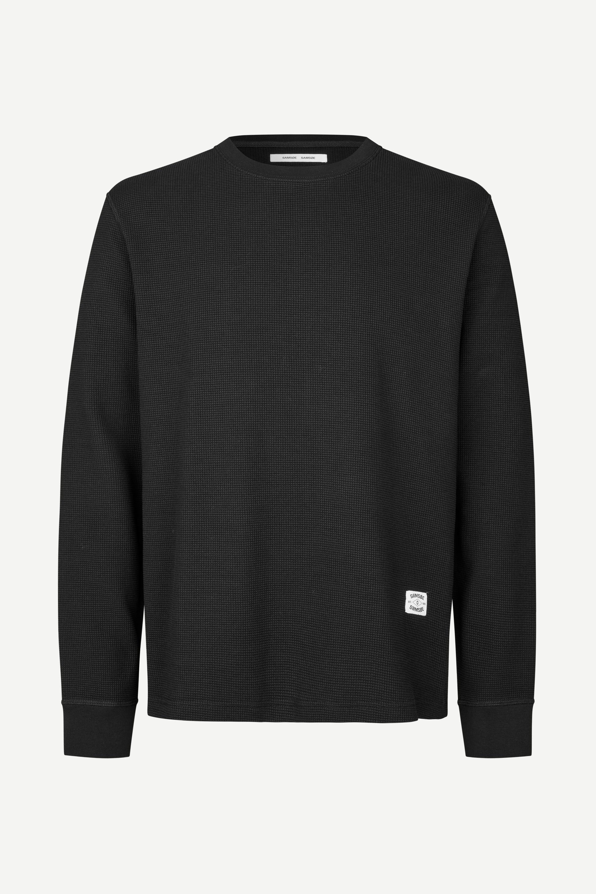 A black long-sleeve crewneck sweatshirt with a small white square logo patch on the lower left hem, displayed on a clean white background.