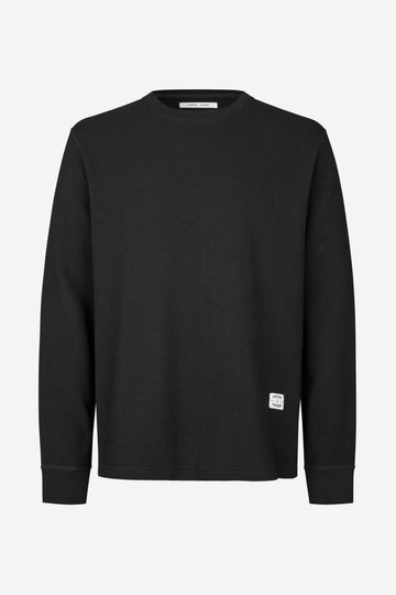 A black long-sleeve crewneck sweatshirt with a small white square logo patch on the lower left hem, displayed on a clean white background.