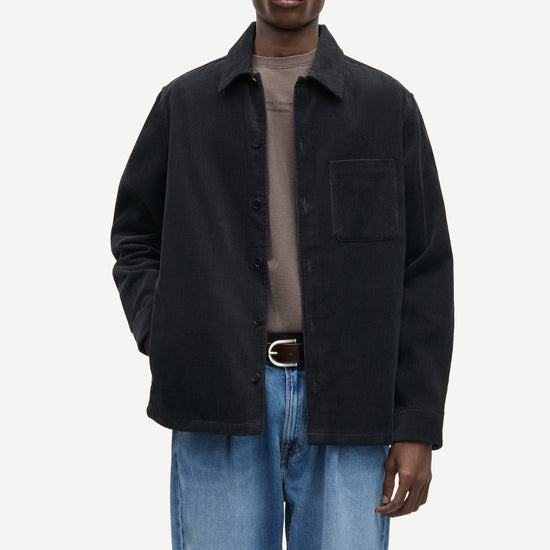 A man modeling a black corduroy overshirt jacket over a taupe t-shirt and blue jeans, photographed against a solid off-white background.