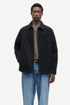 A man modeling a black corduroy overshirt jacket over a taupe t-shirt and blue jeans, photographed against a solid off-white background.