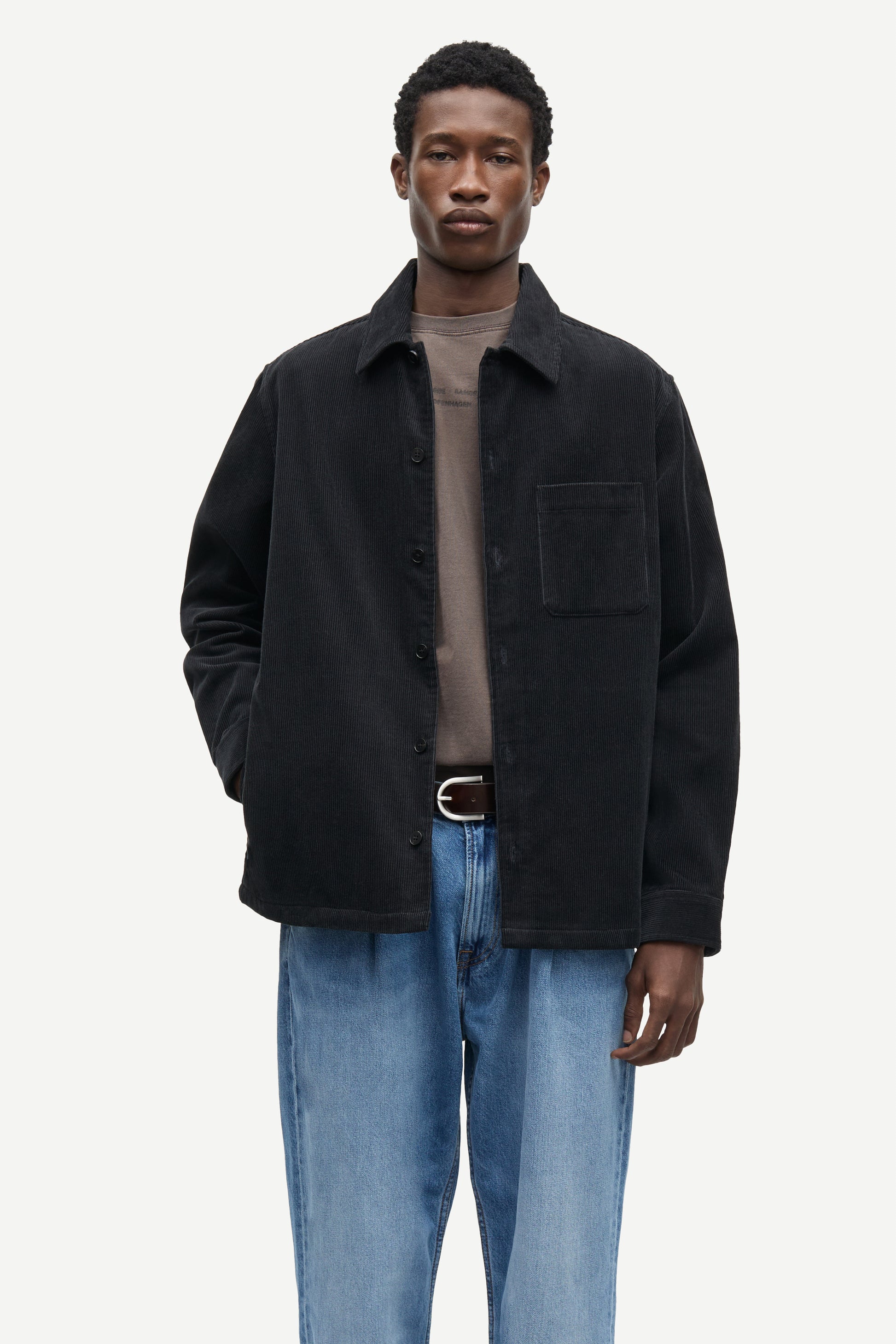 A man modeling a black corduroy overshirt jacket over a taupe t-shirt and blue jeans, photographed against a solid off-white background.