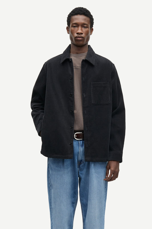 A man modeling a black corduroy overshirt jacket over a taupe t-shirt and blue jeans, photographed against a solid off-white background.