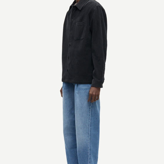 A man wearing a black long-sleeve button-up shirt and light blue jeans, shown in profile against a plain light gray studio background.