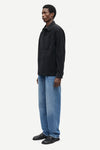 A man wearing a black long-sleeve button-up shirt and light blue jeans, shown in profile against a plain light gray studio background.