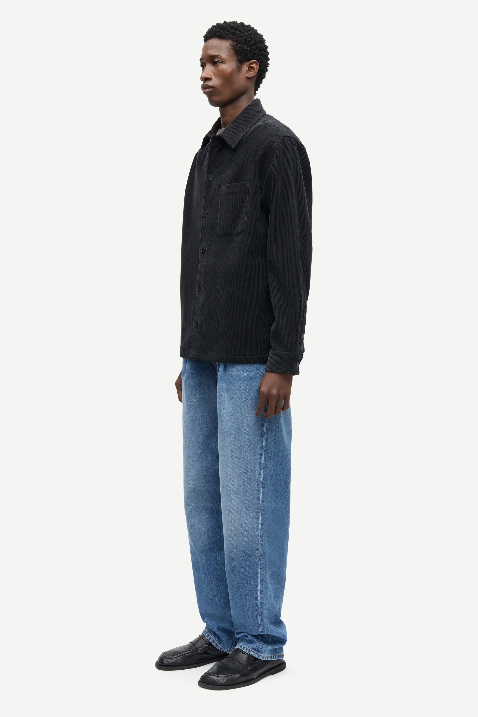 A man wearing a black long-sleeve button-up shirt and light blue jeans, shown in profile against a plain light gray studio background.