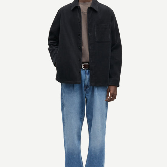 A man modeling a black overshirt jacket over a taupe turtleneck, paired with blue jeans and black leather shoes, against a plain light gray studio background.