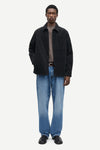 A man modeling a black overshirt jacket over a taupe turtleneck, paired with blue jeans and black leather shoes, against a plain light gray studio background.