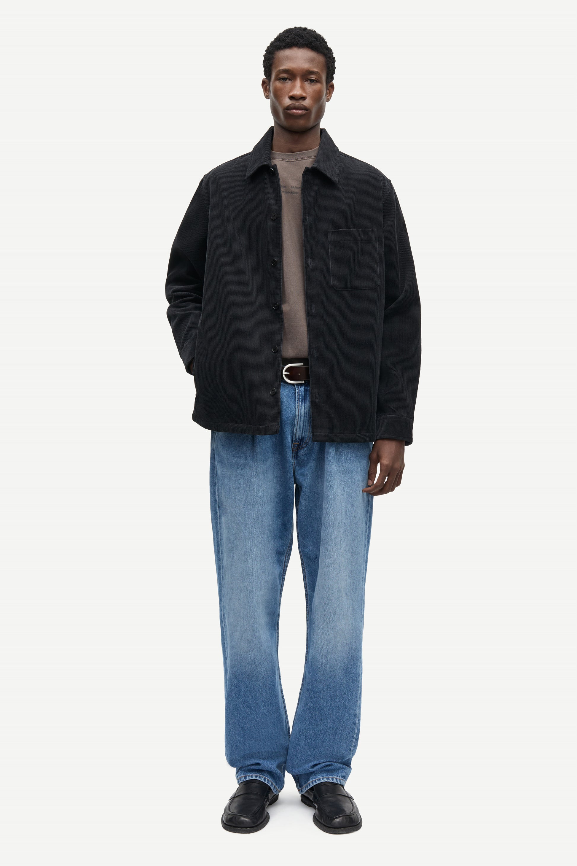 A man modeling a black overshirt jacket over a taupe turtleneck, paired with blue jeans and black leather shoes, against a plain light gray studio background.