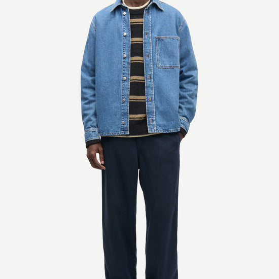 A man modeling a light blue denim overshirt over a black and tan striped shirt, paired with dark navy trousers and brown suede loafers, against a plain light gray background.