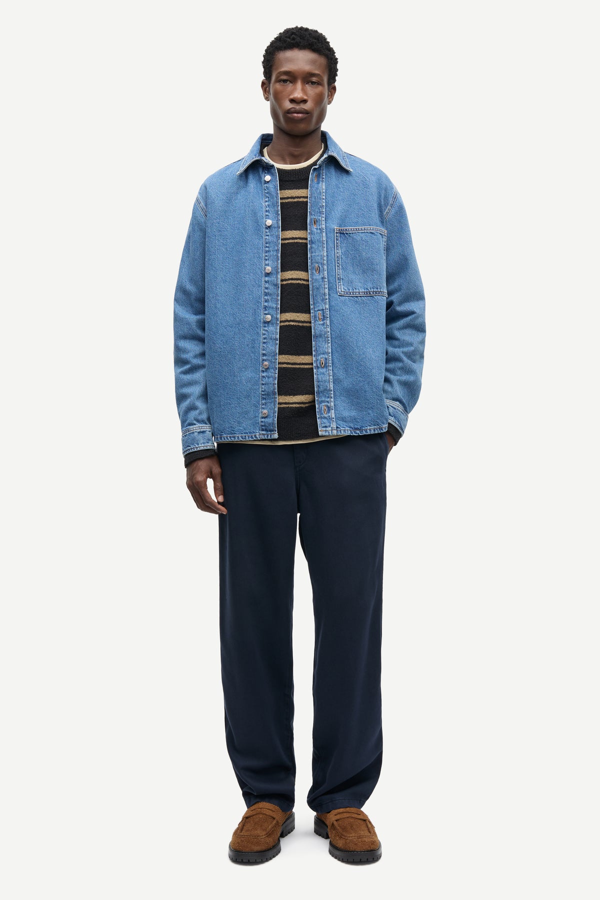 A man modeling a light blue denim overshirt over a black and tan striped shirt, paired with dark navy trousers and brown suede loafers, against a plain light gray background.