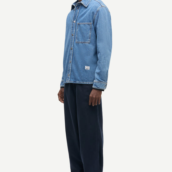 A man wearing a light blue denim button-up shirt, dark navy wide-leg trousers, and brown suede shoes, standing in profile against a light gray studio background.