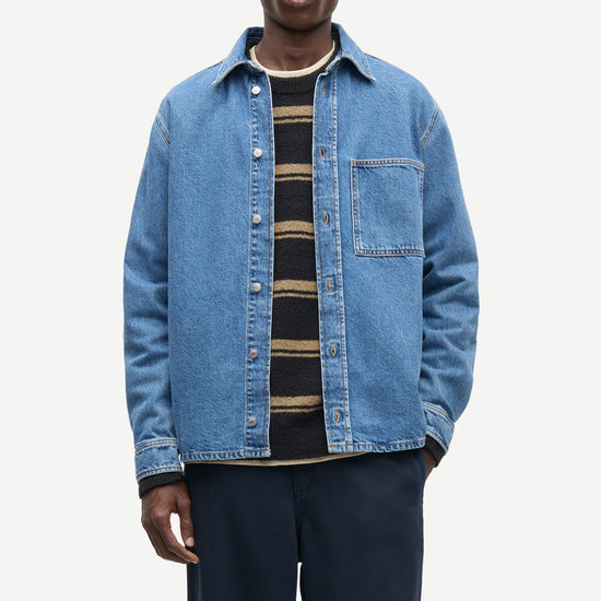 A male model wearing a light blue denim button-up shirt jacket over a black and tan striped knit shirt, paired with dark navy trousers, against a plain light gray background.