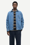A male model wearing a light blue denim button-up shirt jacket over a black and tan striped knit shirt, paired with dark navy trousers, against a plain light gray background.