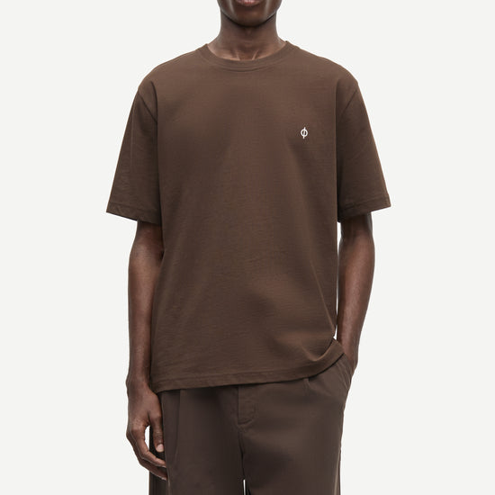 A male model wearing a brown short-sleeve crewneck t-shirt with a small white embroidered logo on the left chest, paired with matching brown trousers, against a light gray studio background.