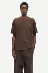 A male model wearing a brown short-sleeve crewneck t-shirt with a small white embroidered logo on the left chest, paired with matching brown trousers, against a light gray studio background.