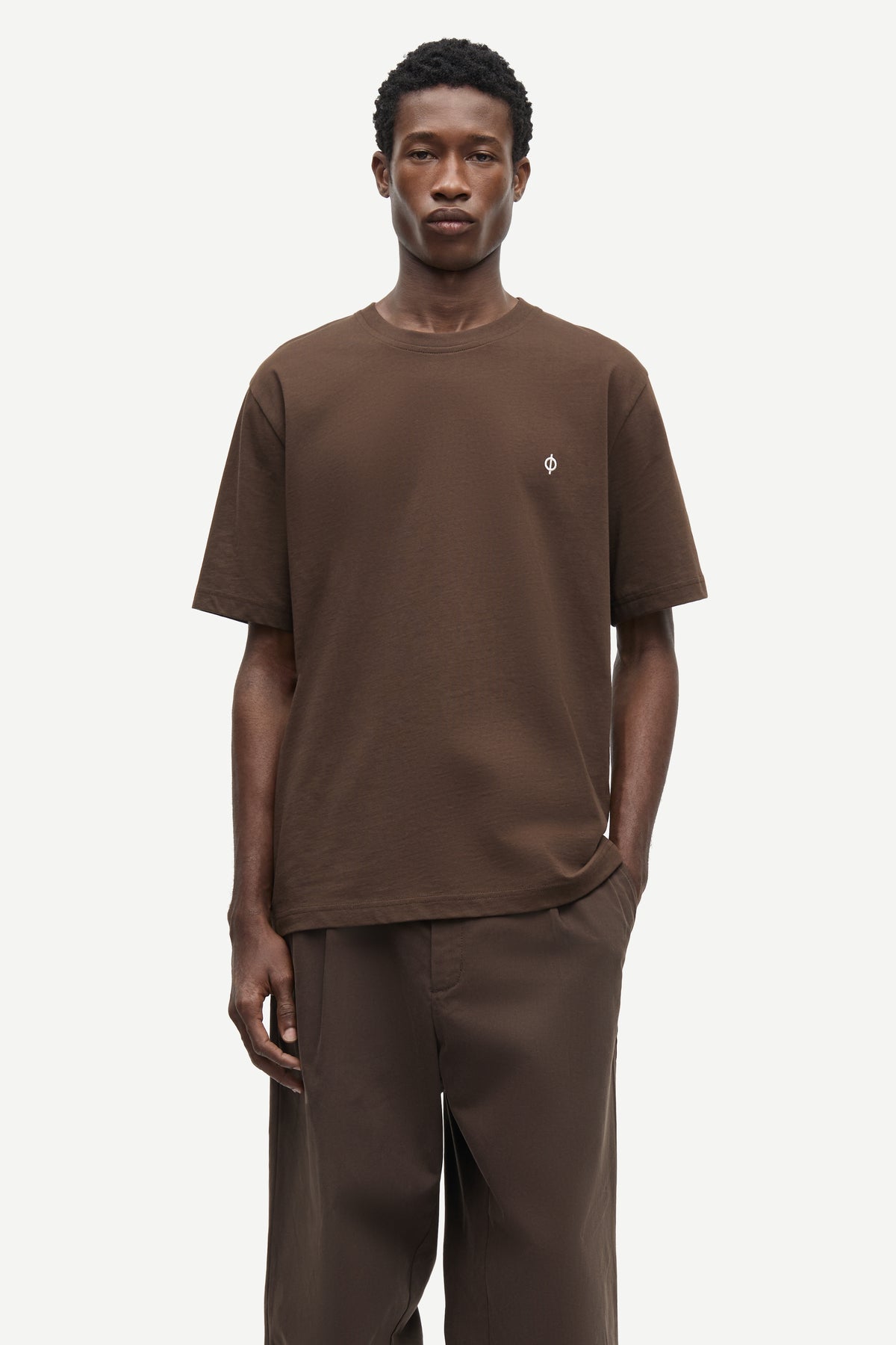 A male model wearing a brown short-sleeve crewneck t-shirt with a small white embroidered logo on the left chest, paired with matching brown trousers, against a light gray studio background.