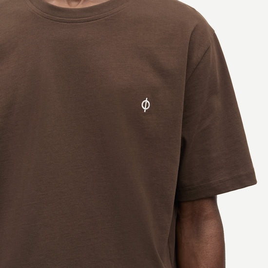 A close-up of a person wearing a brown short-sleeve crewneck t-shirt with a small white circular logo on the left chest, set against a plain light background.