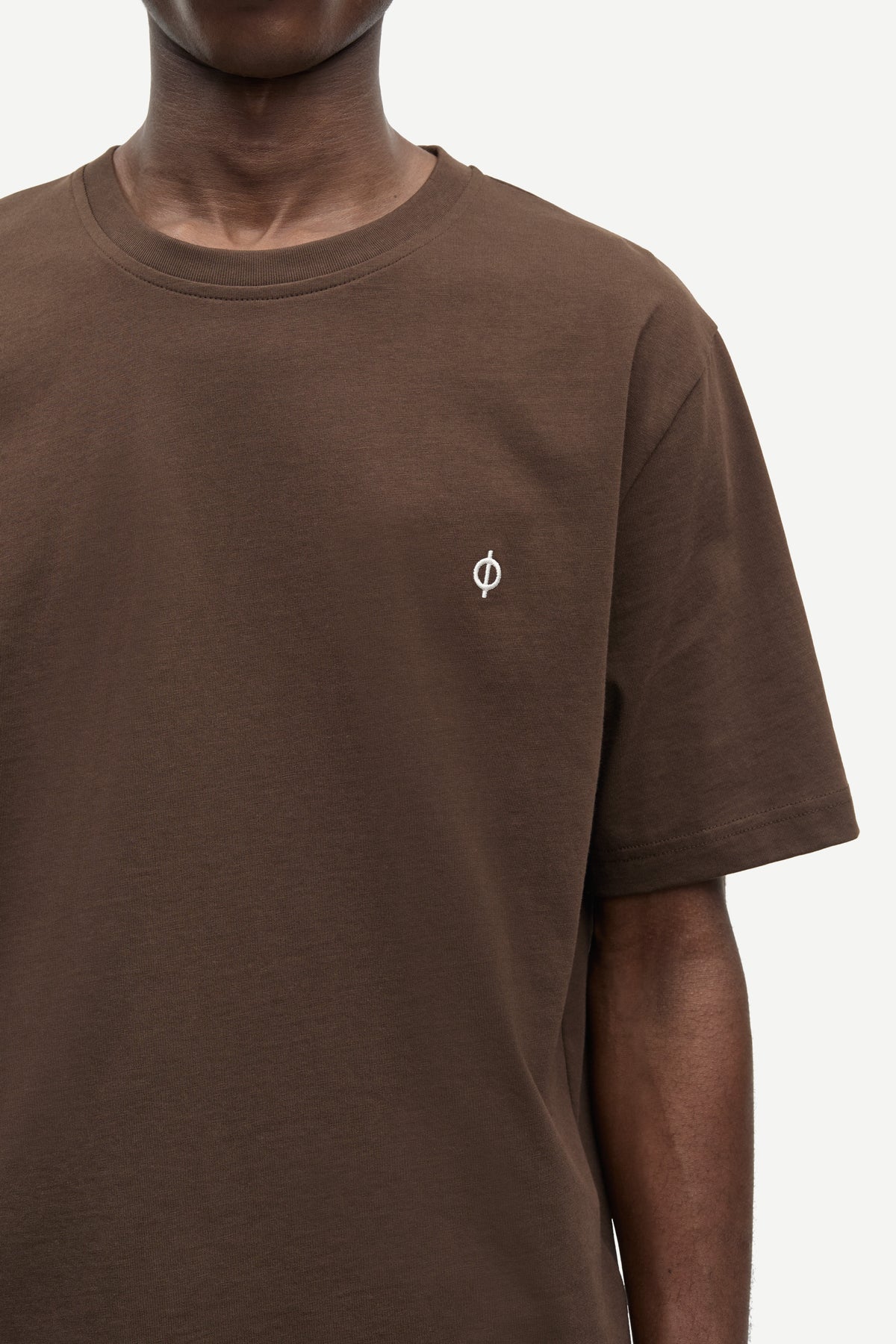 A close-up of a person wearing a brown short-sleeve crewneck t-shirt with a small white circular logo on the left chest, set against a plain light background.