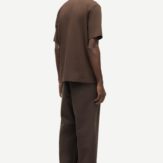 A man viewed from the back wearing a matching brown short-sleeve t-shirt and wide-leg brown trousers against a plain light gray studio background.