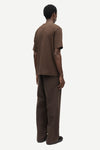 A man viewed from the back wearing a matching brown short-sleeve t-shirt and wide-leg brown trousers against a plain light gray studio background.