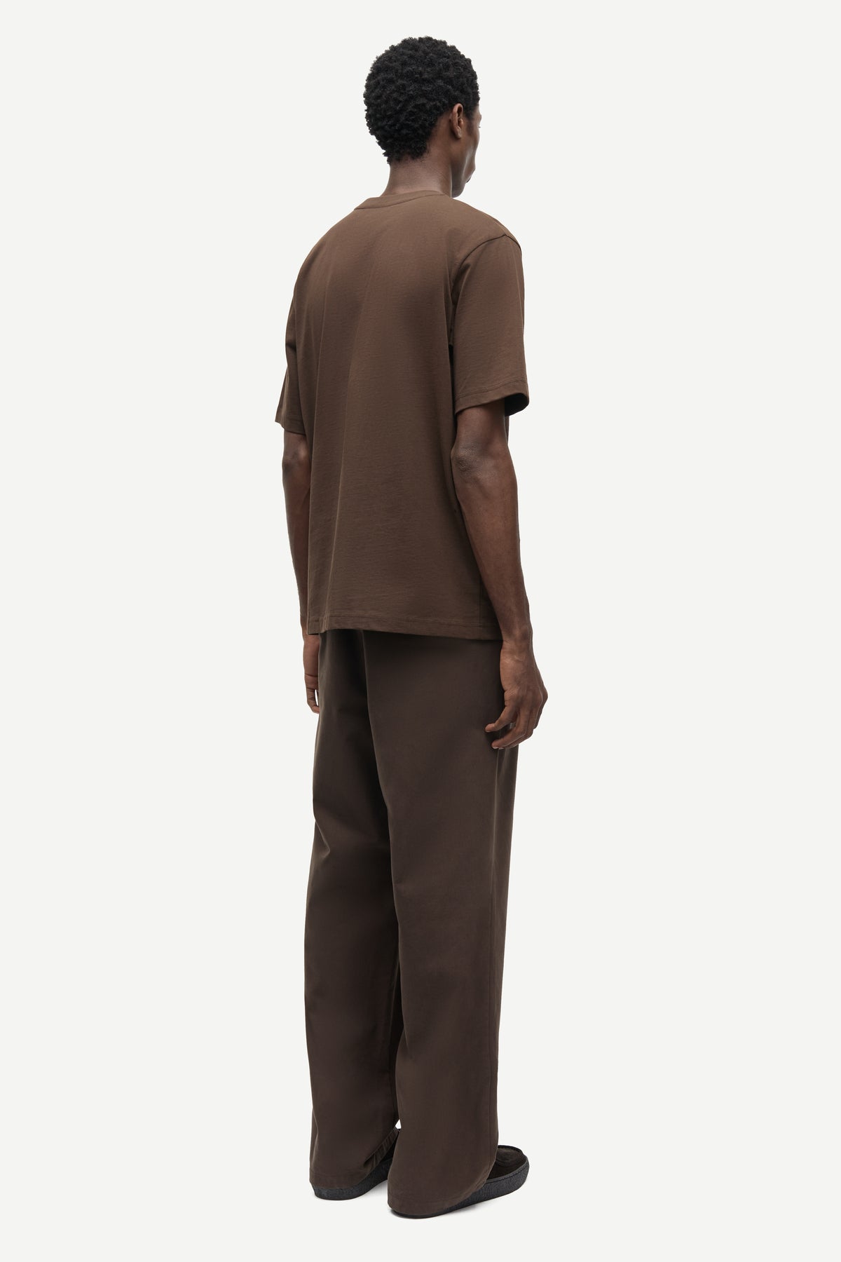 A man viewed from the back wearing a matching brown short-sleeve t-shirt and wide-leg brown trousers against a plain light gray studio background.