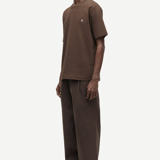 A man wearing a brown short-sleeve t-shirt and matching brown wide-leg trousers, standing in profile against a light gray studio background.