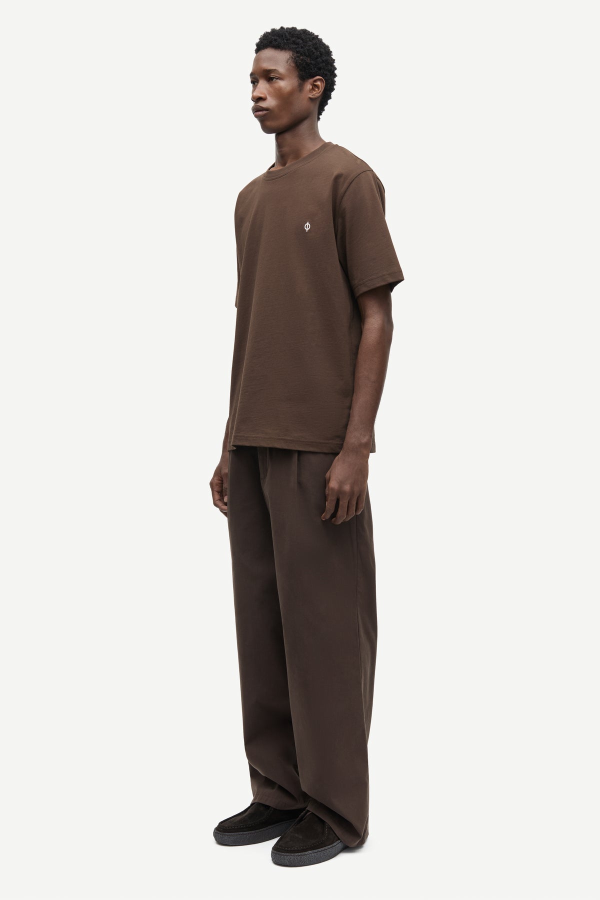 A man wearing a brown short-sleeve t-shirt and matching brown wide-leg trousers, standing in profile against a light gray studio background.
