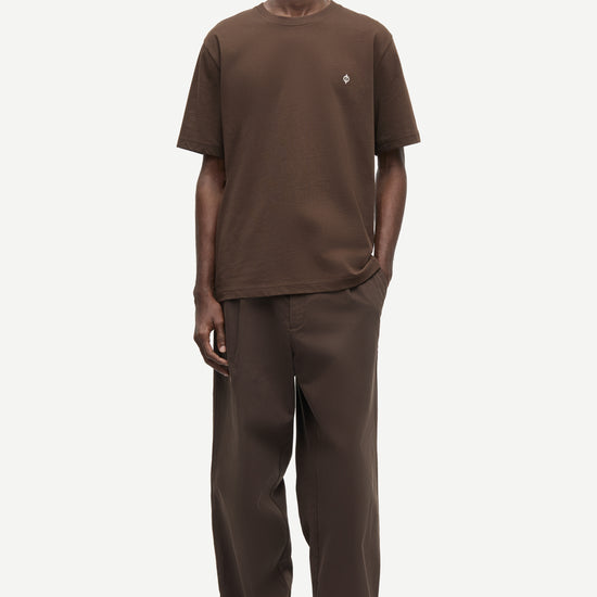 A man modeling a matching brown short-sleeve crewneck t-shirt and wide-leg brown trousers against a light gray studio background.