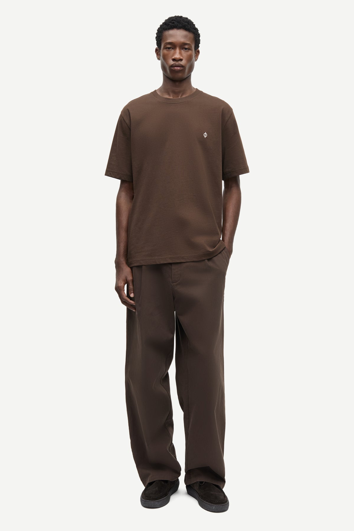 A man modeling a matching brown short-sleeve crewneck t-shirt and wide-leg brown trousers against a light gray studio background.