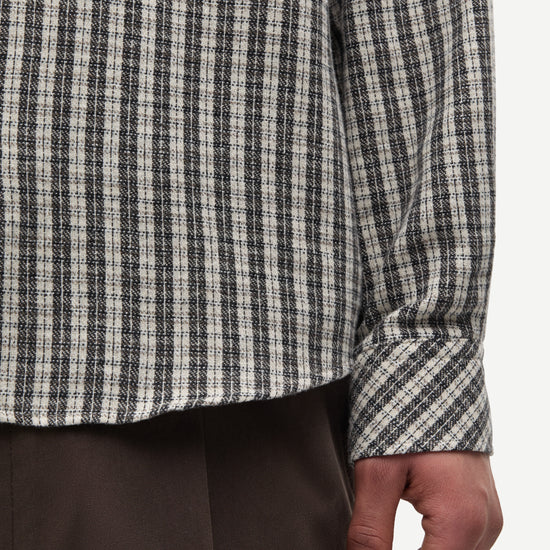 Close-up of a person wearing a long-sleeve plaid shirt in black, white, and gray, paired with dark brown trousers, against a plain light background.