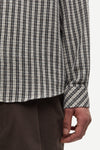 Close-up of a person wearing a long-sleeve plaid shirt in black, white, and gray, paired with dark brown trousers, against a plain light background.